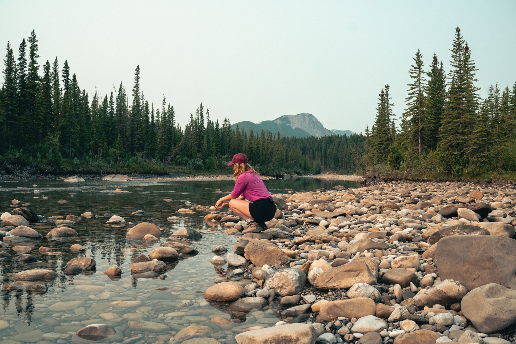 A person sitting beside a rocky river with forest and mountains beyond