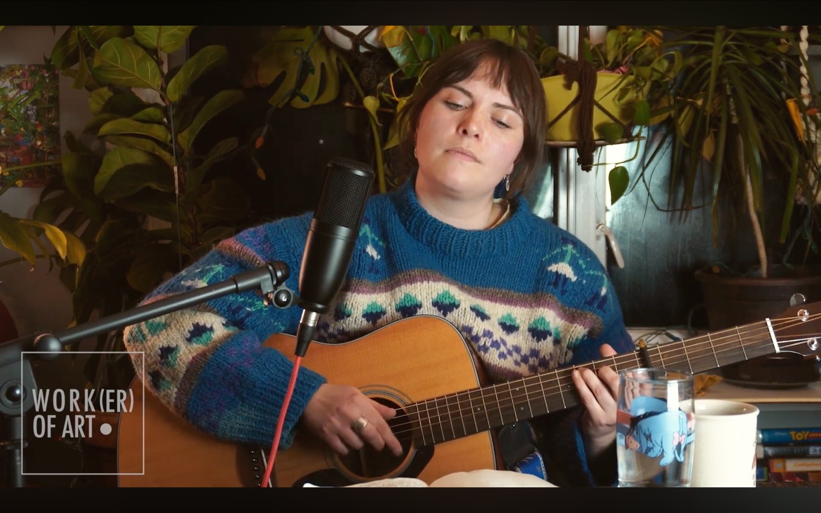 A musician performing with a guitar and microphone in a Worker of Art artist feature